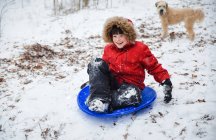 Menino feliz escorregando por uma colina no dia de inverno nevado, enquanto o cão assiste. — Fotografia de Stock