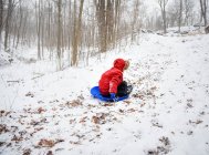 Junge in rotem Mantel rodelt an einem verschneiten Wintertag im Wald den Berg hinunter. — Stockfoto