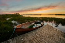 Barco de madera dañado con agua y muelle de madera ubicado cerca de la costa cubierta de hierba y lago tranquilo contra el cielo nublado al atardecer - foto de stock