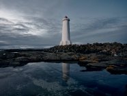 Lighthouse tower located on rocky cliff near calm reflective sea water against gray overcast sky — Stock Photo