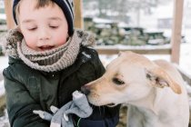 Petit garçon jouant dans la neige dehors avec le chiot curieux à côté de lui — Photo de stock