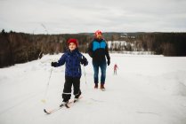 Garçon amusant d'hiver souriant sur les skis avec la famille derrière lui le jour neigeux — Photo de stock