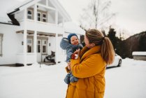 Мама і чарівна дитина посміхається в холодний сніжний день біля білого будинку — стокове фото