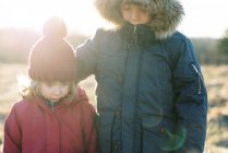 Двое детей стоят на вершине холма в зимней одежде и смотрят на закат — стоковое фото