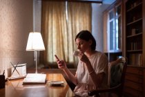 Junge Frau blättert im Smartphone und schlürft Heißgetränk, während sie Buch liest — Stockfoto