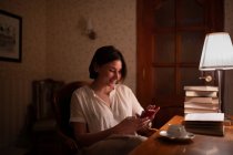 Glückliche junge Frau lächelt und surft mit dem Handy, während sie abends Bücher liest — Stockfoto