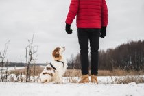 O cão feliz confiante com os olhos fechados senta-se ao lado de seu proprietário ao ar livre por um rio nevado congelado. Caminhadas, caminhadas com animais de estimação no inverno - o homem de casaco de inverno vermelho desfruta de tempo livre com seu cão — Fotografia de Stock