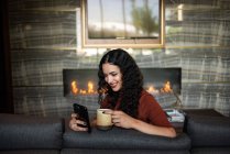 Jeune femme avec tasse de café et téléphone portable à la maison — Photo de stock