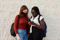 Duas jovens mulheres com uma máscara estão olhando para seu smartphone. — Fotografia de Stock