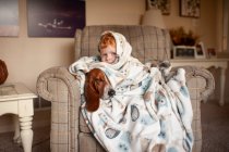 Boy 3-4 years old cuddles with dog in large blanket sitting in chair — Stock Photo