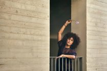 Belle jeune fille aux cheveux afro tient une fleur. Elle se penche sur un balcon. — Photo de stock