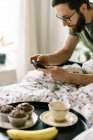 Мужчина завтракает на кровати дома и с помощью смартфона — стоковое фото