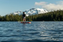 Anônimo feminino caminhante paddleboarding no lago perto da encosta da montanha e floresta verde — Fotografia de Stock