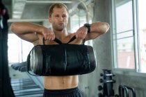 Homem forte exercitar levantando sacos de peso, homem de fitness muscular fazendo agachamentos usando saco de fitness no ginásio. — Fotografia de Stock