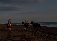 Hombres a caballo en la playa al atardecer - foto de stock