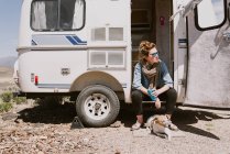 A stylish young woman and her dog sit outside their car trailer — Stock Photo