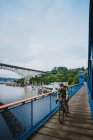 Un jeune homme fait du vélo sur un pont avec de l'eau et des bateaux au loin — Photo de stock