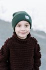 Kleiner lächelnder Junge mit Hut im hausgemachten Pullover, der draußen steht — Stockfoto