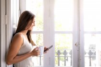 Mujer joven usando el teléfono móvil mientras está sentada en el alféizar de la ventana en casa - foto de stock