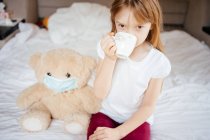 Девушка пьет чай с плюшевым мишкой в медицинской маске на белой кровати — стоковое фото