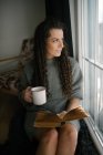 Красива молода жінка читає книгу біля вікна в затишному домашньому інтер'єрі — стокове фото