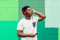 Niño negro afroamericano sobre fondo de pared verde. Escuchar música con auriculares y teléfono móvil. - foto de stock
