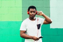 Afro ragazzo nero americano su sfondo muro verde. Ascolto di musica con cuffie e telefono cellulare. — Foto stock