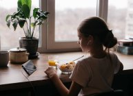 A menina toma café da manhã e olha para o telefone — Fotografia de Stock