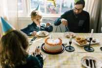 Una pequeña familia comiendo pastel de cumpleaños en la mesa y celebrando - foto de stock