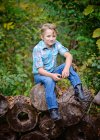 Jovem loiro de olhos azuis menino sentado em uma pilha de madeira no país. — Fotografia de Stock