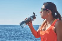 Latin woman, middle-aged, resting, regaining strength, eating, drinking water, after a gym session, burning calories, keeping fit, outdoors by the sea, wearing headphones and smart watch — Stock Photo