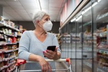 Donna anziana caucasica con maschera facciale e negozio di telefoni nel supermercato — Foto stock