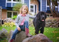 Jovem loira menina sentado fora no um rocha com Grande preto cão — Fotografia de Stock