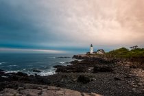Morning blue hour colors fill the sky while off in the distance, the Portland Head Lighthouse shines its light. — Stock Photo