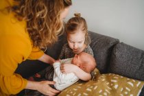 Mom helping older daughter to hold newborn baby on sofa at home — Stock Photo