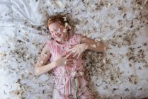 Young red haired girl laying on the bed during a feather pillow fight. — Stock Photo