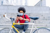 Junger Mann mit Afro-Haaren sitzt neben seinem alten Fahrrad und sendet eine Nachricht — Stockfoto