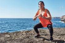 Latin woman, middle-aged, with orange top, black leggings, training, doing physical exercises, squats, burning calories, keeping fit, outdoors by the sea, blue sky, day, sunny winter, bluetoo — Stock Photo