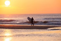 A surfer walking along the beach at sunset — Stock Photo