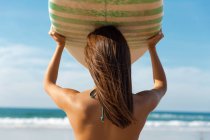 Schönes Surfermädchen mit einem Surfbrett über dem Kopf, während es auf die Wellen blickt — Stockfoto