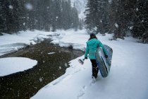 Задний вид женщины, несущей доску во время прогулки по снежному полю — стоковое фото