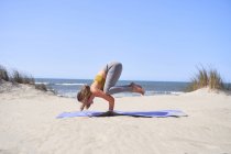 Chica joven realizando Bakasana yoga posición en la playa por la mañana. Meditación y concepto de vida saludable. - foto de stock