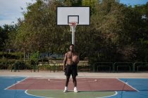 Fuerte jugador afroamericano con pelota descansando cerca de aro de baloncesto en la cancha - foto de stock
