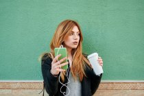Donna d'affari in possesso di una tazza di caffè mentre guarda il suo telefono ascoltando musica su uno sfondo verde — Foto stock