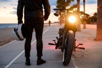 Jeune homme debout près de la moto et profitant de la vue sur le coucher du soleil — Photo de stock