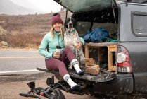 Mujer joven sonriente sentada con el perro en el maletero del vehículo todoterreno durante la nevada - foto de stock