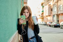 Mujer de negocios sosteniendo una taza de café mientras mira su teléfono escuchando música sobre un fondo verde - foto de stock