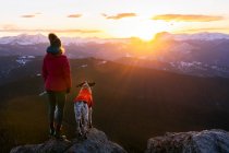 Женщина с собакой смотрит на пейзаж с вершины горы во время заката — стоковое фото
