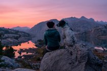 Вид спереду молодої жінки, що сидить на скелі з собакою на горі під час заходу сонця — стокове фото