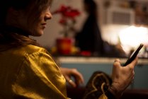 Mujer en camisa de seda de oro en casa acogedora cocina mira el dispositivo del teléfono - foto de stock